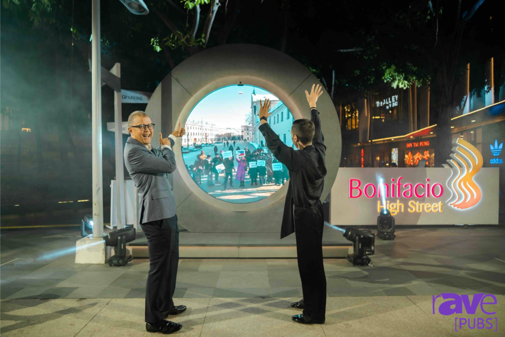 Two people stand in front of a city portal running Video Window, gesturing towards an animated scene displayed inside it. Colorful lights and signage are visible in the background, creating a lively atmosphere.