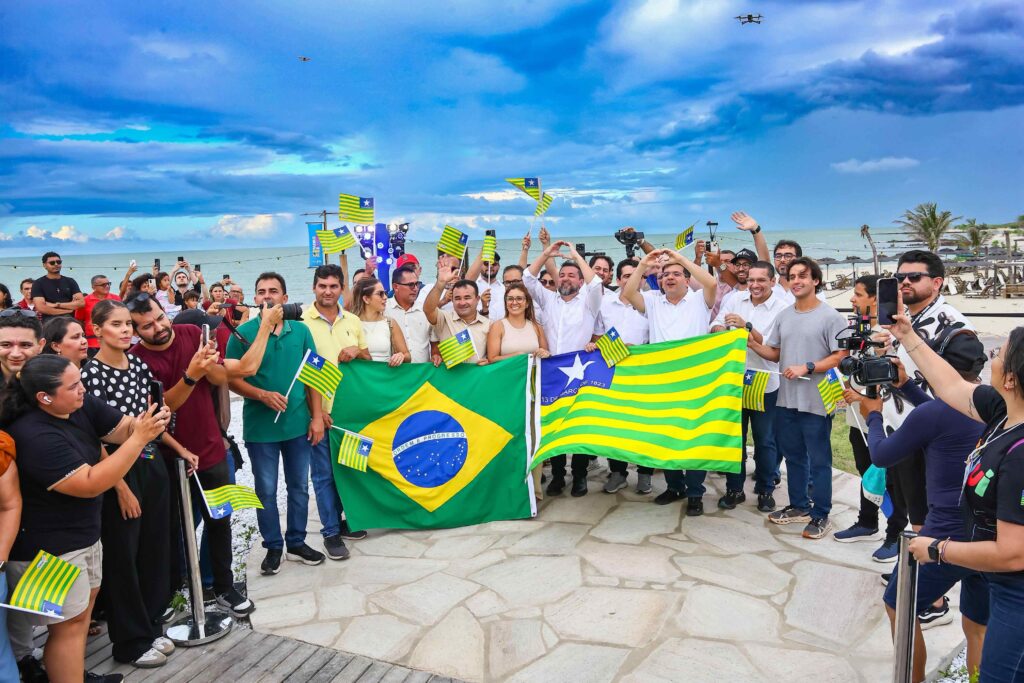 A diverse group of people gathers outdoors, joyfully waving Brazilian flags and celebrating the launch of the Brazilian Portal in Piauí. The backdrop features a cloudy sky and a coastal view.