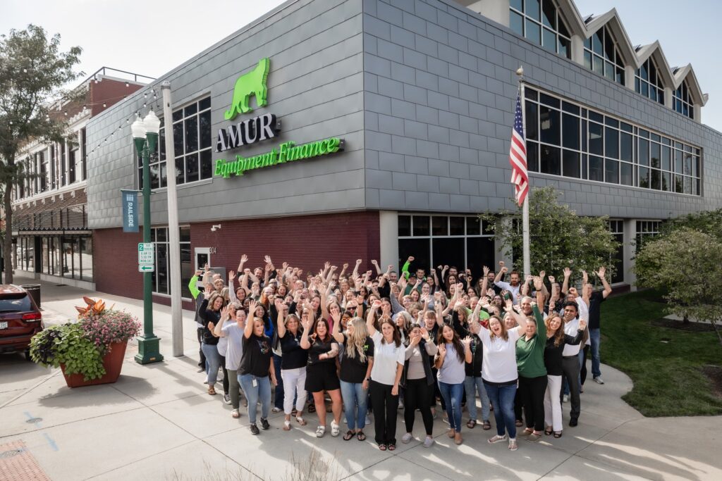 A large group of people stands outside a modern building with a green sign that reads "AMUR" and an American flag nearby. The crowd is smiling and raising their hands in celebration.